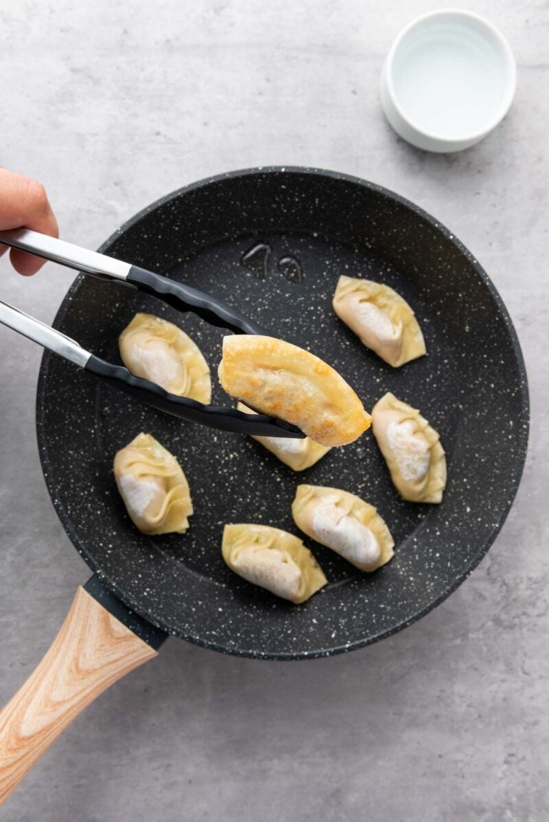 A hand uses tongs to pick up a dumpling from a pan showing the bottom crispy surface.