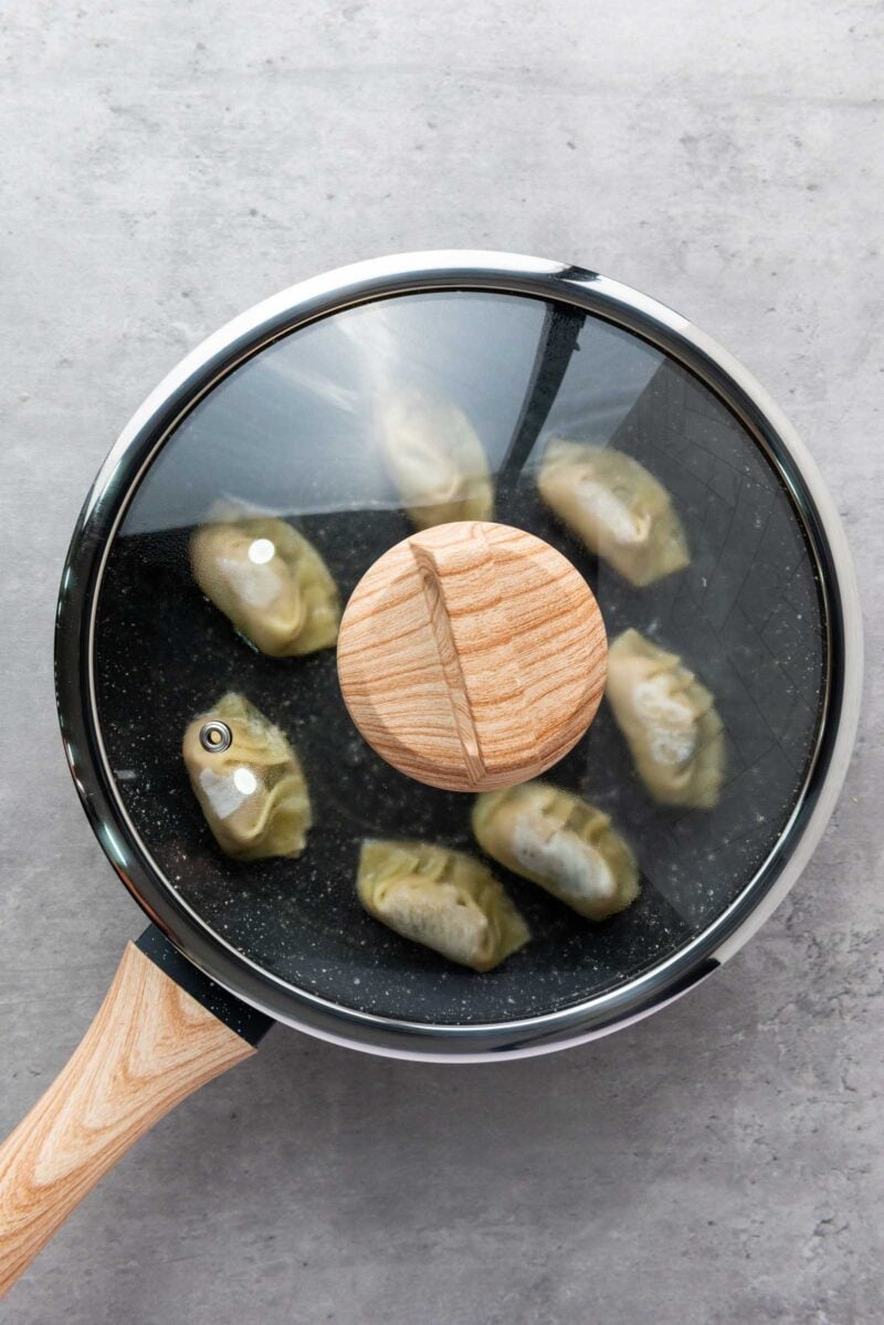 A frying pan with a glass lid contains dumplings steaming.
