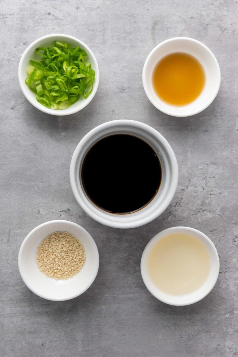 Five small bowls on a gray surface, each containing green onions, soy sauce, rice vinegar, sesame seeds, and sesame oil.