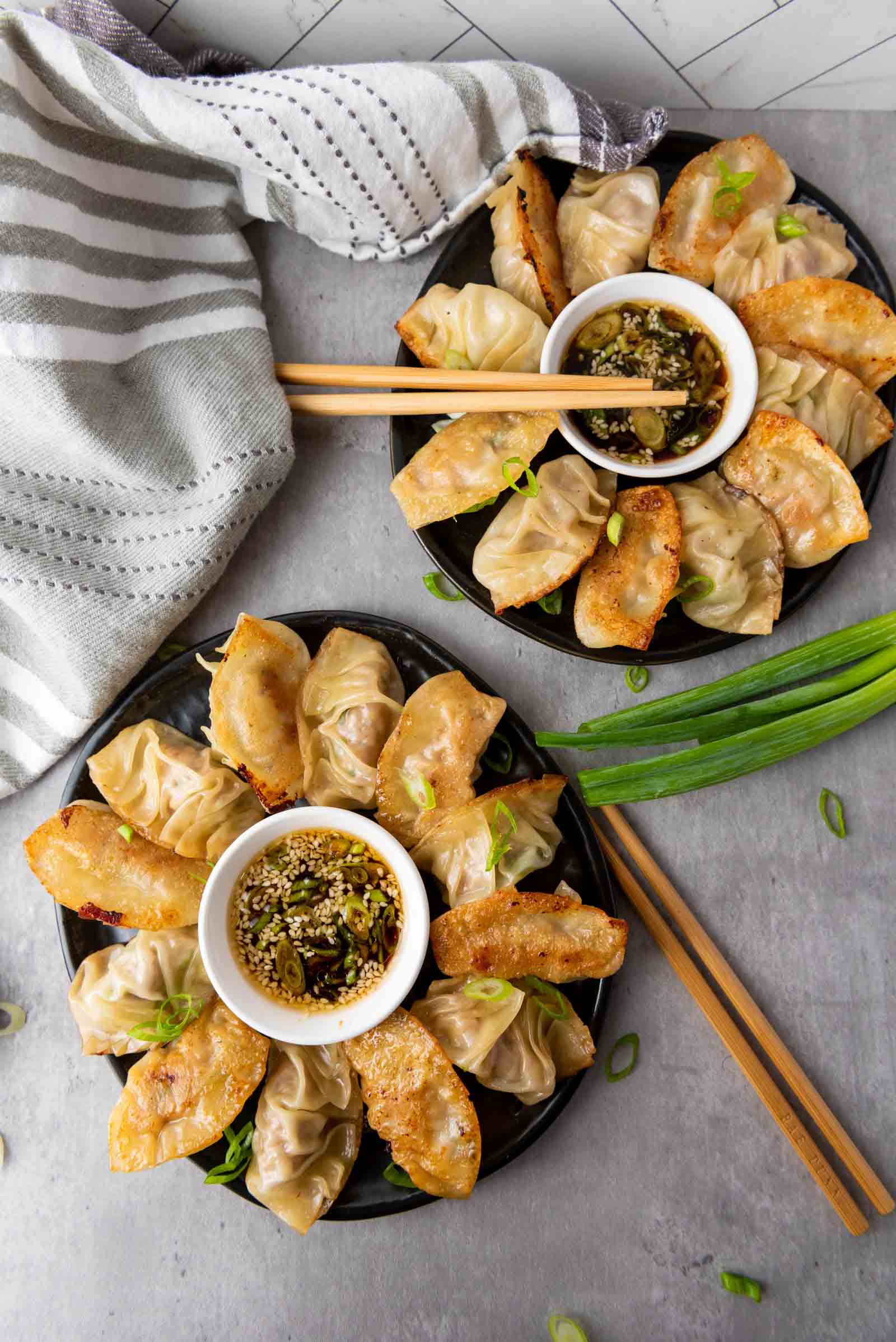 Two plates of pan-fried pork dumplings arranged in a circle around dipping sauce, garnished with green onions, with chopsticks and a striped towel on a gray surface.