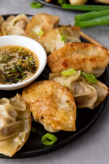 A black plate with pan-fried dumplings, garnished with green onions, next to a small bowl of soy dipping sauce topped with sesame seeds and sliced scallions.