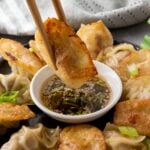 A hand uses chopsticks to dip a pan-fried dumpling into a small bowl of dipping sauce, surrounded by more dumplings on a plate.