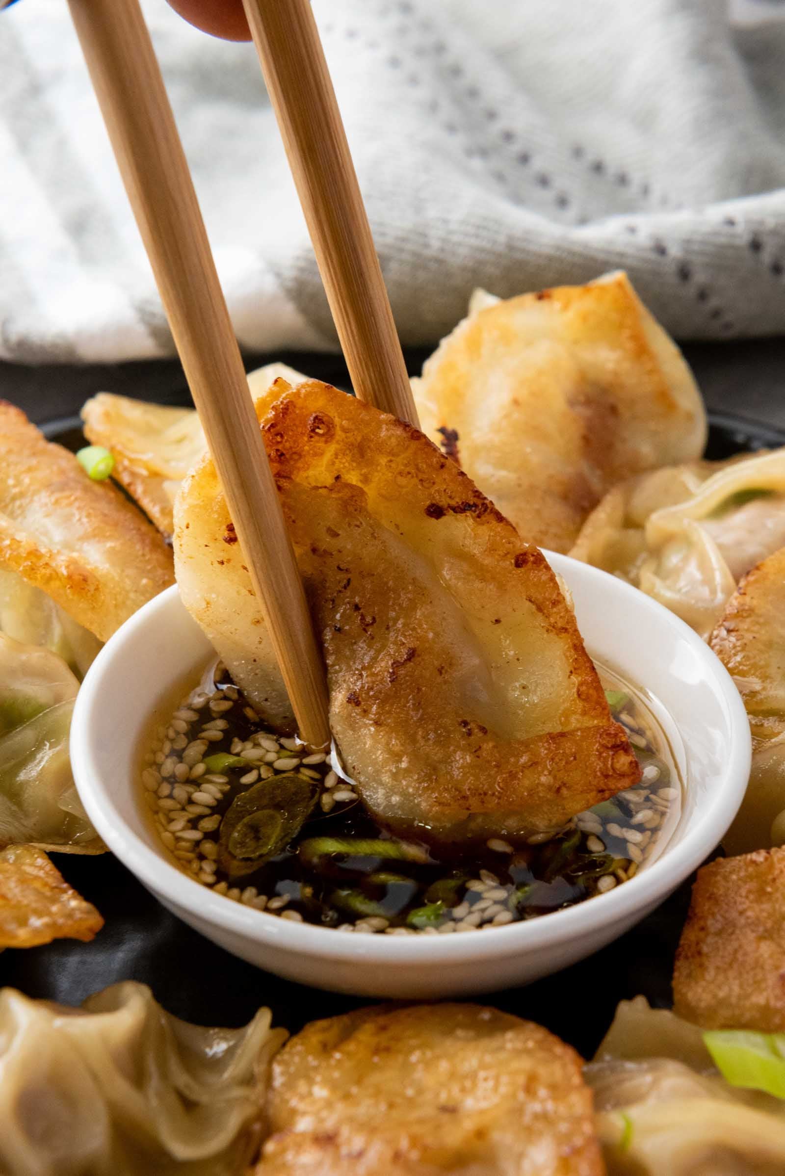 A fried dumpling is picked up with chopsticks and dipped into a small bowl of sauce with sesame seeds and green onions, surrounded by more dumplings.