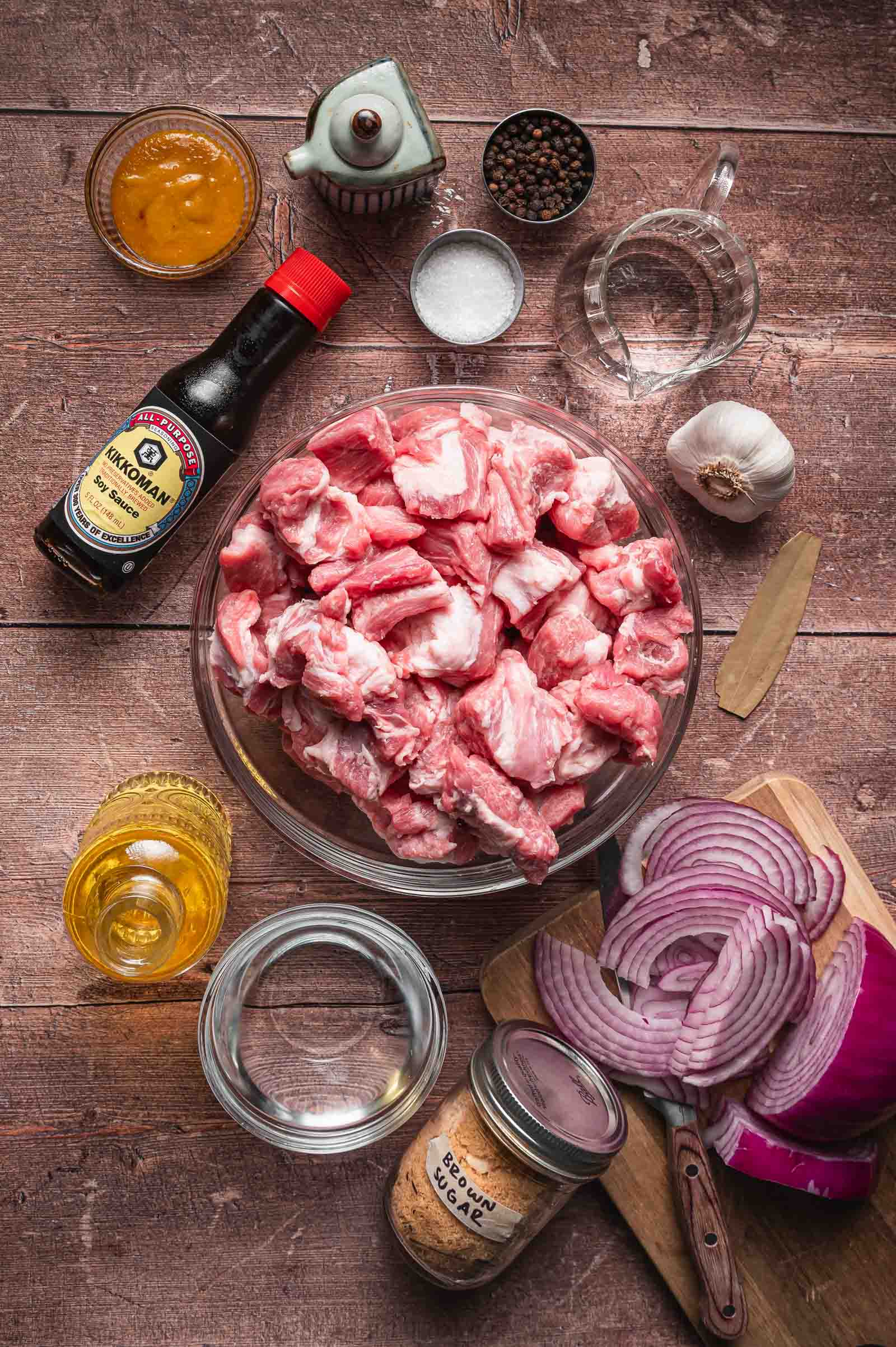 Top-down view of raw pork chunks in a bowl surrounded by soy sauce, oil, spices, garlic, vinegar, sliced red onion, and seasonings on a wooden surface.