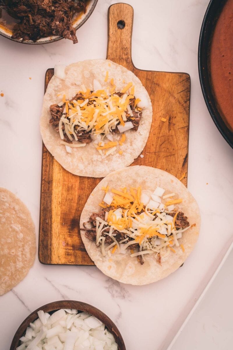Two corn tortillas topped with shredded beef, chopped onions, and shredded cheese sit on a wooden board. Nearby are a bowl of diced onions, a tortilla, and a pan with red sauce.