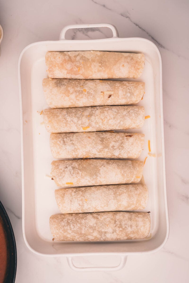 Beef filled and rolled tortillas are arranged in a white rectangular baking dish on a marble surface.