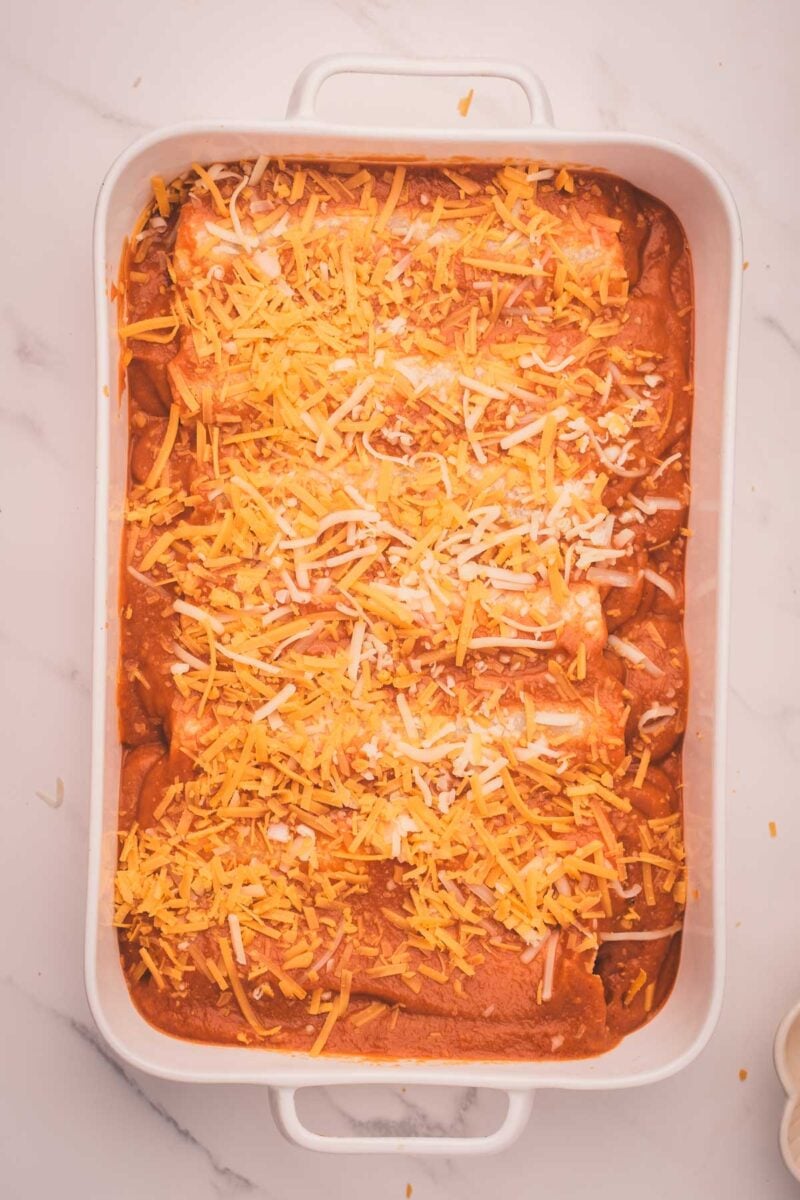 A white baking dish filled with enchiladas topped with red sauce and shredded cheddar cheese on a marble surface.