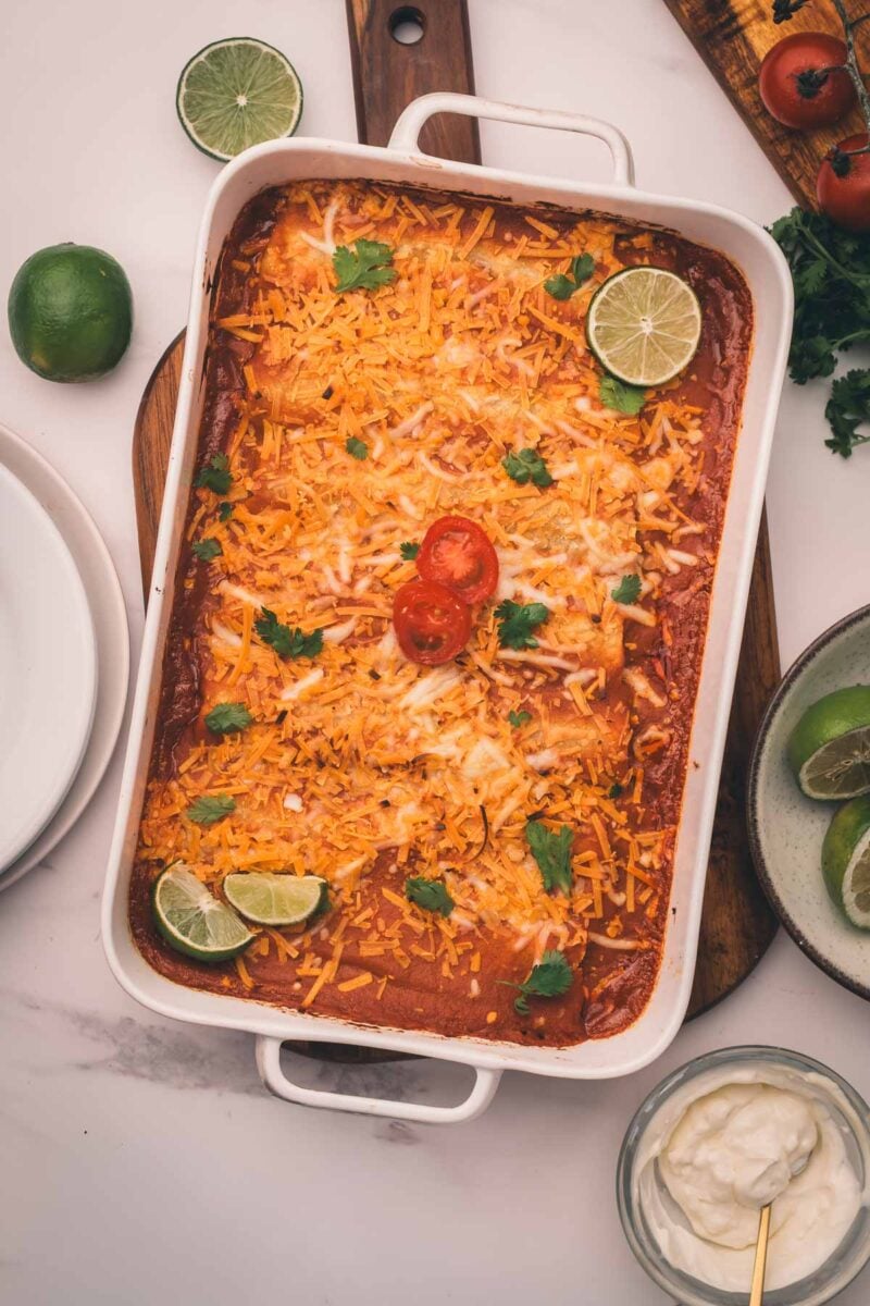 A casserole dish filled with baked enchiladas, topped with melted cheese, cilantro, lime wedges, and sliced cherry tomatoes. Plates, limes, and a bowl of sour cream are nearby.
