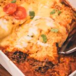 A close-up of shredded beef enchiladas in a white baking dish topped with melted cheese, sliced tomato, lime, and cilantro, with a serving spatula lifting one enchilada.