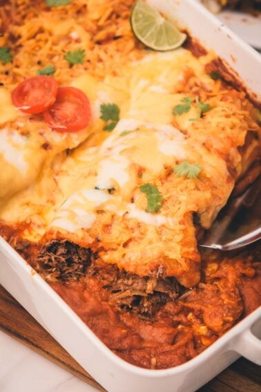 A close-up of shredded beef enchiladas in a white baking dish topped with melted cheese, sliced tomato, lime, and cilantro, with a serving spatula lifting one enchilada.