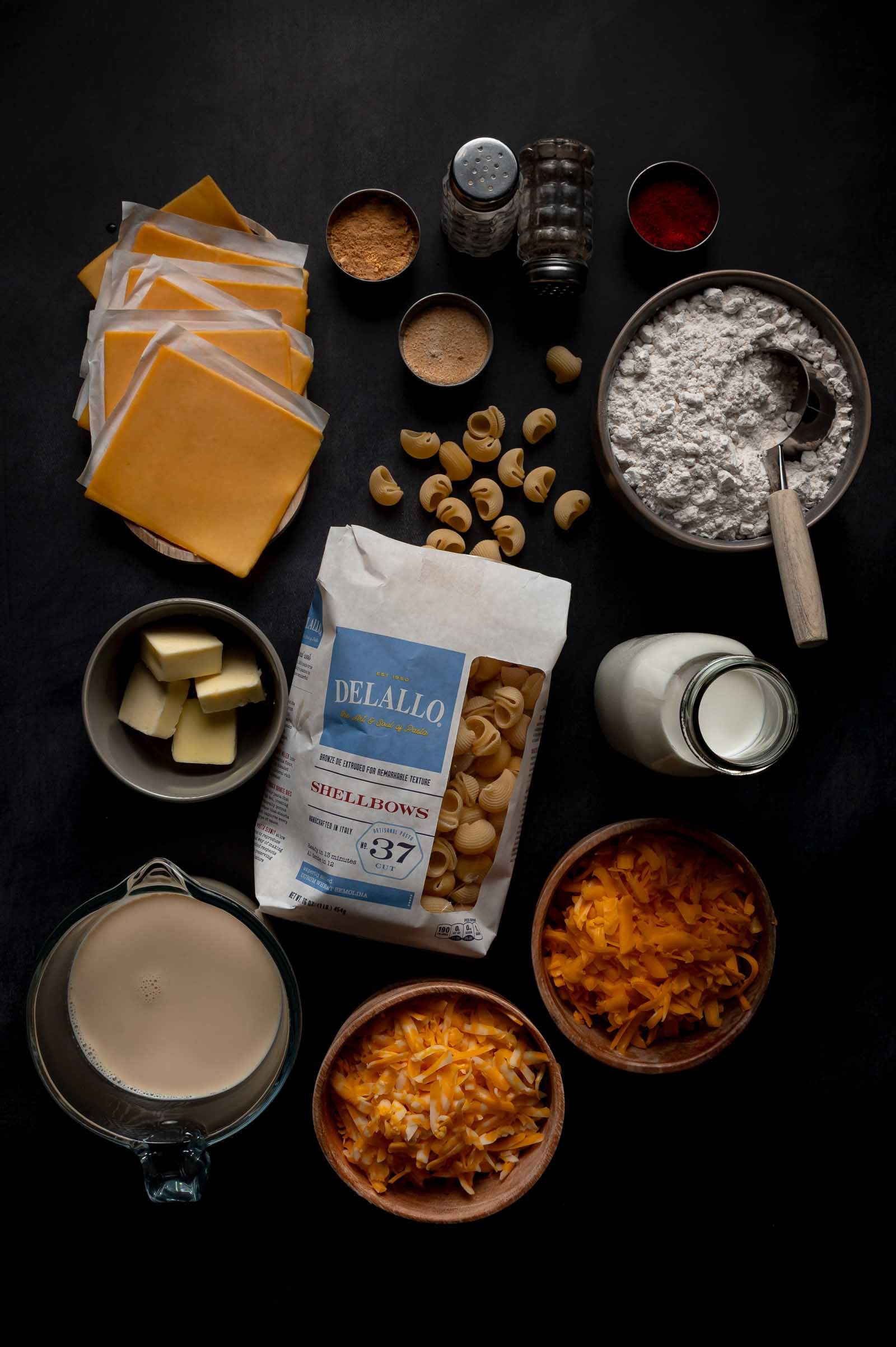 Overhead view of ingredients for mac and cheese, including pasta, cheese slices, shredded cheese, flour, butter, milk, seasonings, and a measuring cup of liquid on a dark surface.