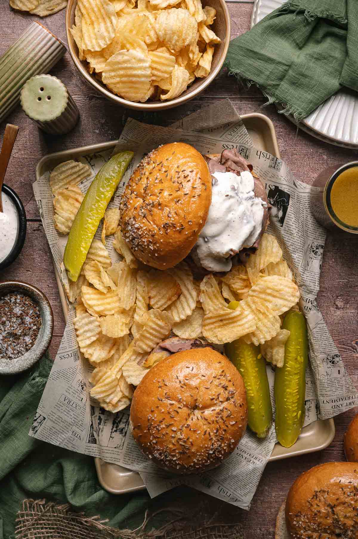 Two beef on weck sandwiches with sauce are served on a tray with wavy potato chips and pickle spears, placed over newspaper-style paper.