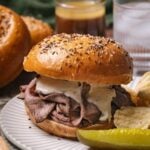 A beef on weck sandwich with creamy sauce on a salted and seeded bun, served with potato chips and a pickle spear on a white plate.