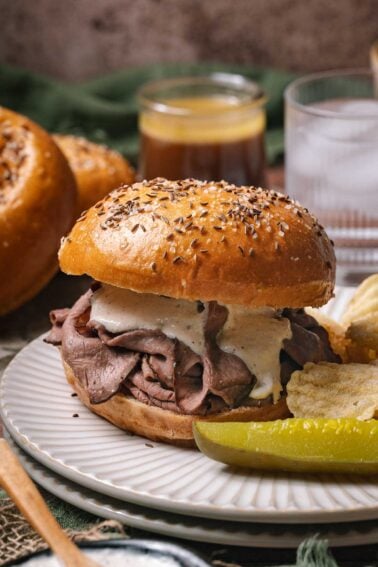 A beef on weck sandwich with creamy sauce on a salted and seeded bun, served with potato chips and a pickle spear on a white plate.