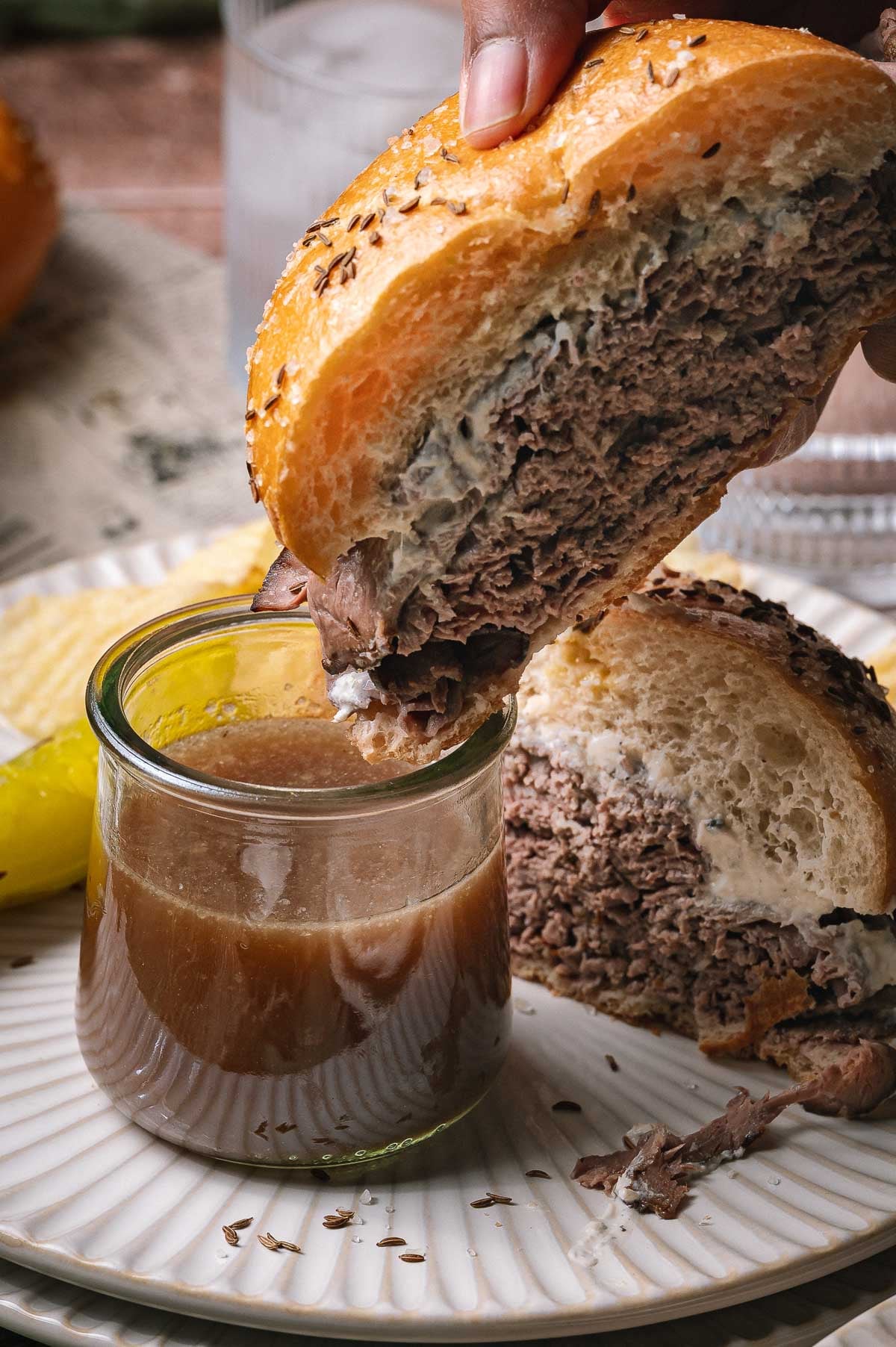A hand dips a beef on weck with a salted bun into a small jar of au jus sauce on a plate with chips and a pickle.