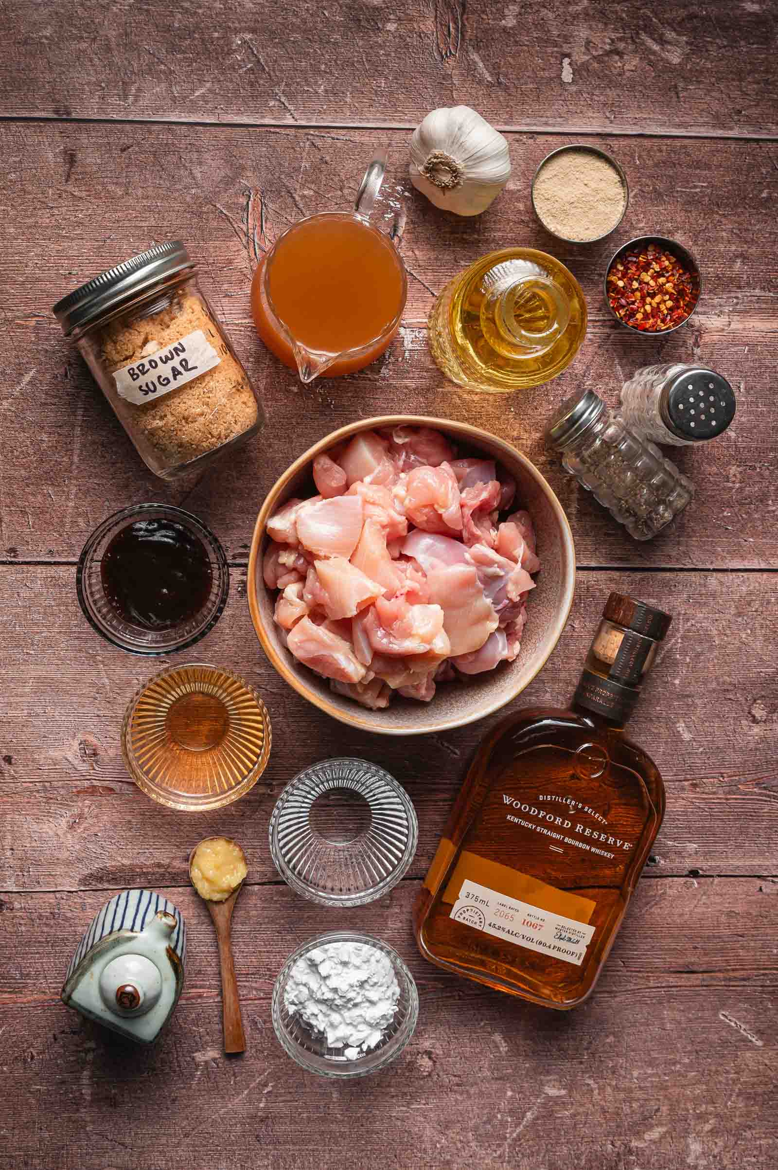 Overhead view of raw diced chicken in a bowl surrounded by brown sugar, bourbon, oil, garlic, soy sauce, cornstarch, seasonings, and broth on a wooden surface.