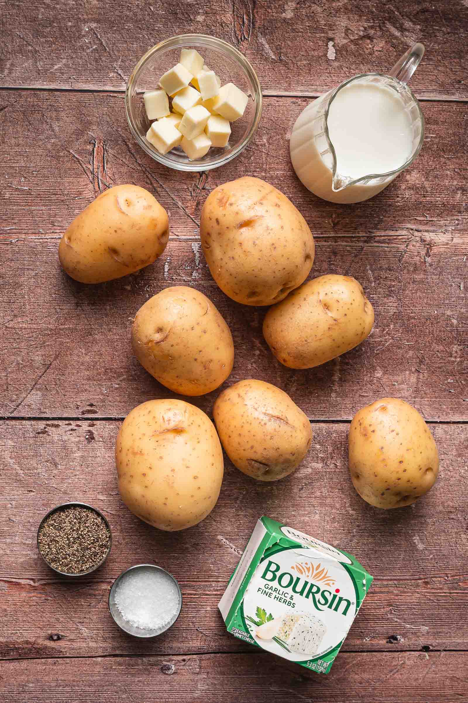 Ingredients on a wooden surface: six Yukon Gold potatoes, butter cubes in a bowl, milk in a glass pitcher, black pepper, salt, and a package of Boursin cheese.