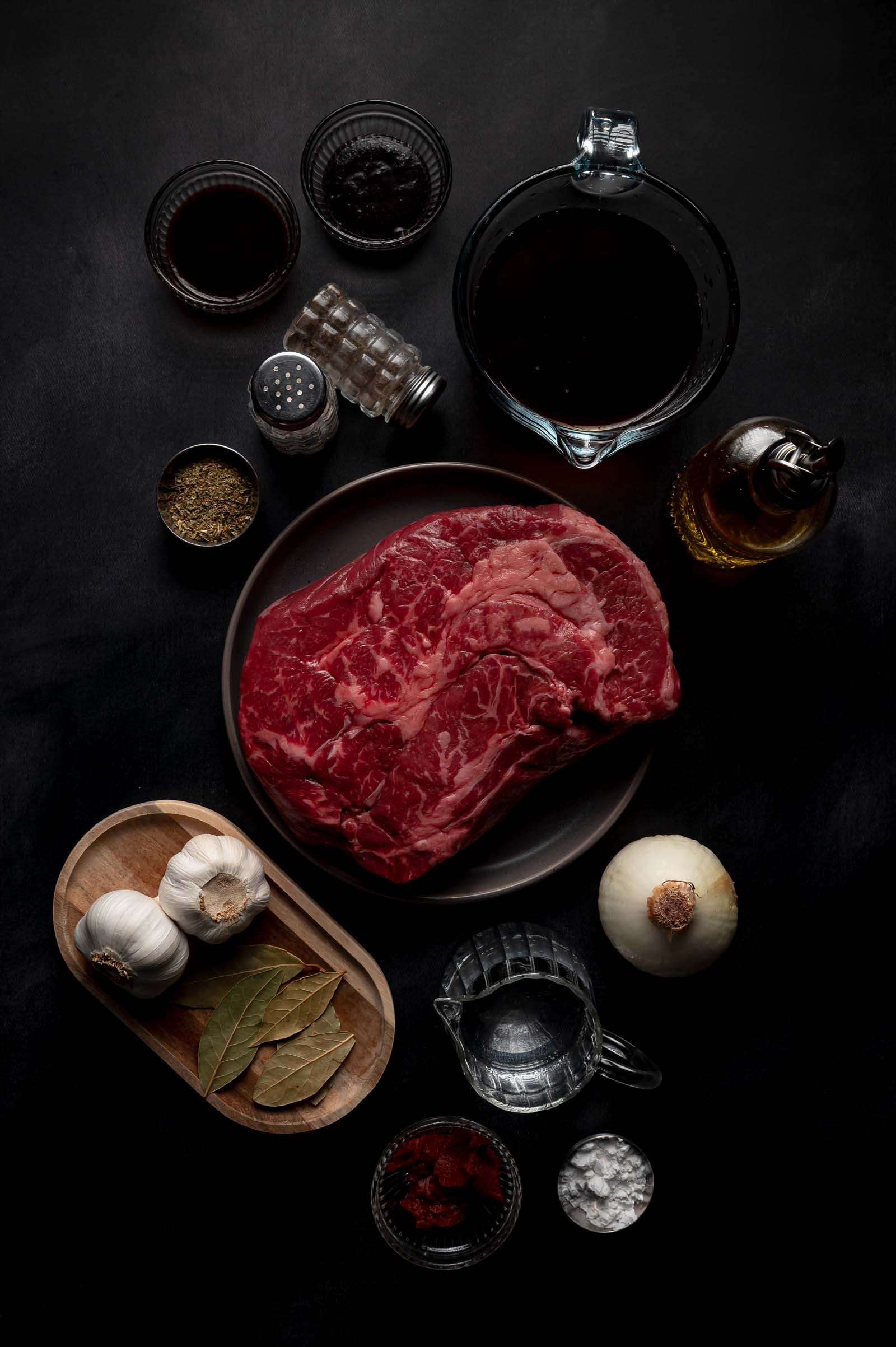 Raw beef surrounded by various ingredients including garlic, bay leaves, whole onion, oil, spices, soy sauce, vinegar, and seasonings, all arranged on a dark surface.
