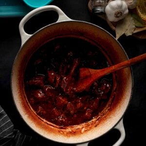 A Dutch oven with beef tips in gravy, stirred by a wooden spoon. Garlic, bay leaves, salt, and pepper shakers are nearby on a dark surface.