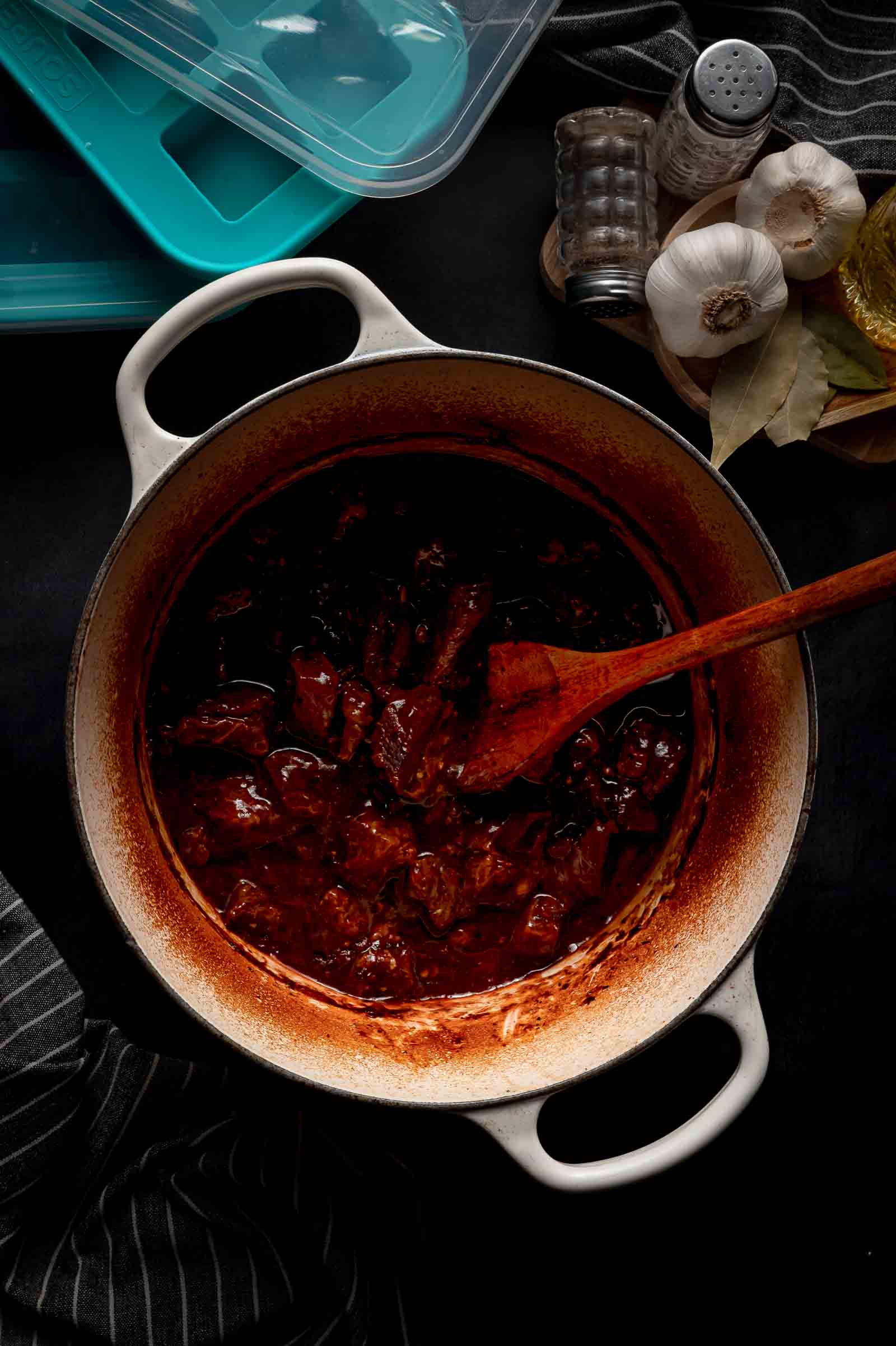 A Dutch oven with beef tips in gravy, stirred by a wooden spoon. Garlic, bay leaves, salt, and pepper shakers are nearby on a dark surface.