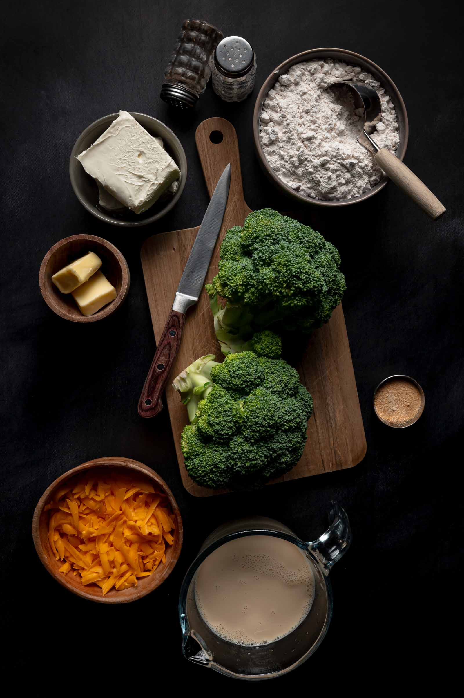 Overhead view of broccoli, shredded cheese, butter, cream cheese, flour, milk, spices, and a knife arranged on a dark surface.