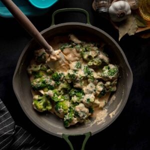 Broccoli florets coated in a creamy cheese sauce are being stirred in a deep pan; nearby are seasonings, garlic, bay leaves, and storage containers.