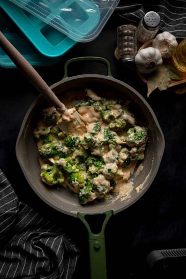 Broccoli florets coated in a creamy cheese sauce are being stirred in a deep pan; nearby are seasonings, garlic, bay leaves, and storage containers.
