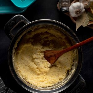 A pot of cream cheese mashed potatoes with a wooden spoon rests on a dark surface, next to garlic bulbs, bay leaves, salt, and pepper shakers.