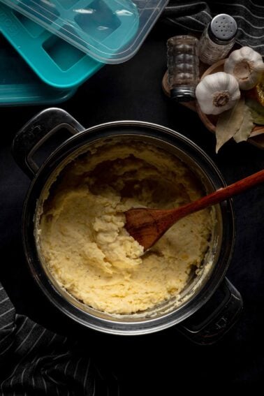 A pot of cream cheese mashed potatoes with a wooden spoon rests on a dark surface, next to garlic bulbs, bay leaves, salt, and pepper shakers.