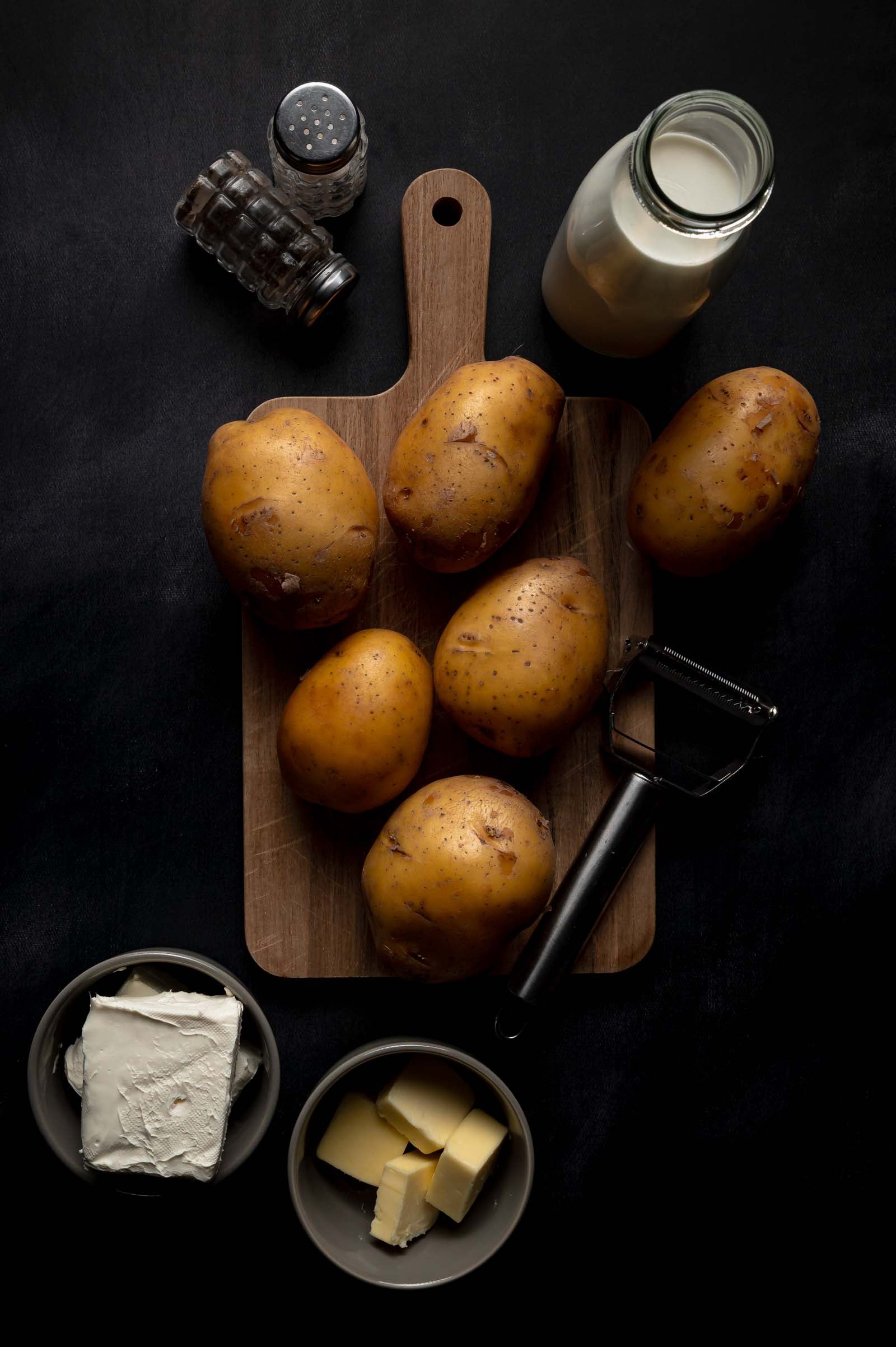 Whole potatoes on a wooden board with a peeler, surrounded by milk, butter, cream cheese, salt, and pepper on a dark surface.