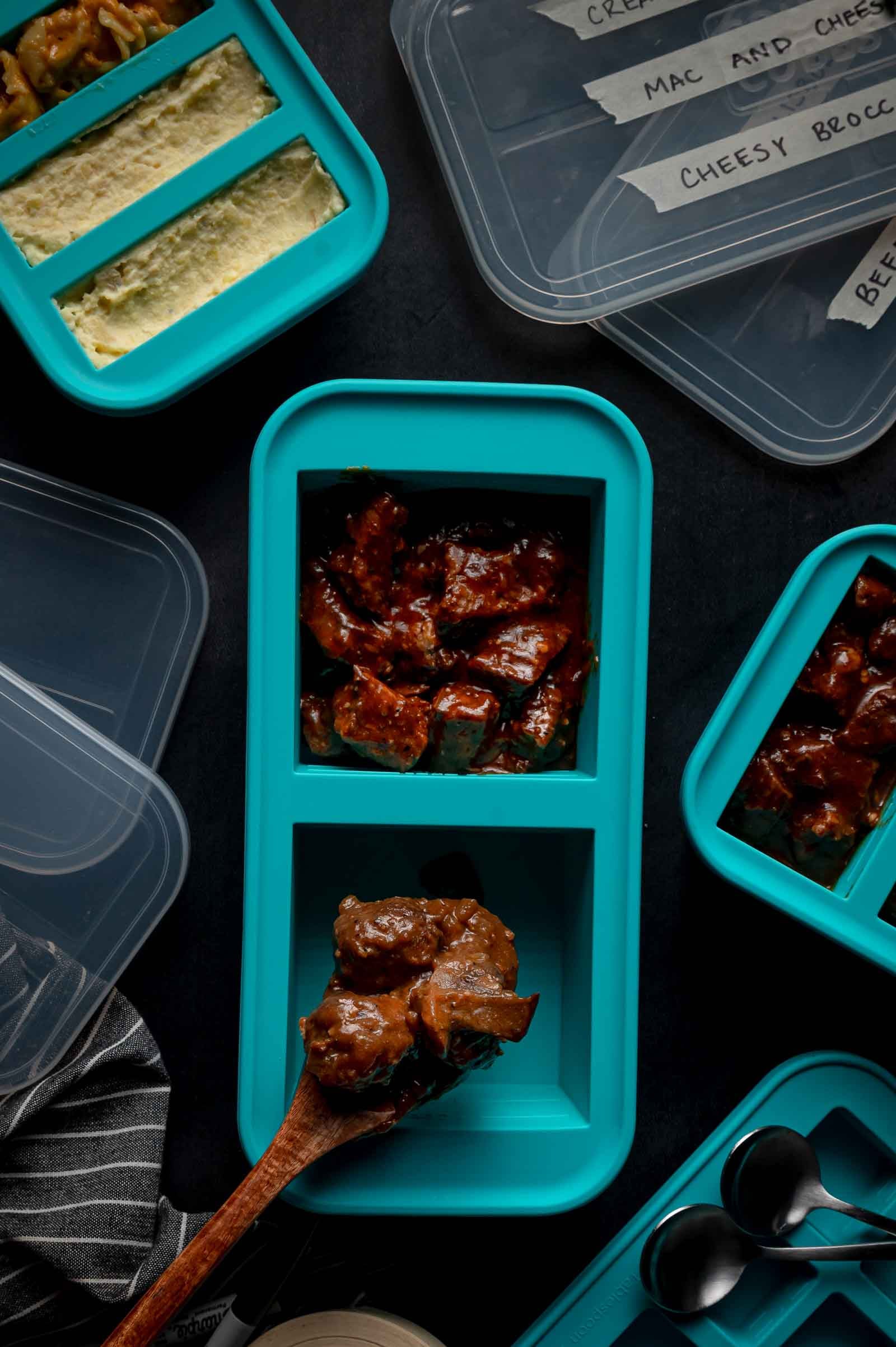 Blue meal prep containers filled with food, including beef tips in one slot, with labeled lids nearby on a dark surface. A wooden spoon is scooping meatballs into one compartment.