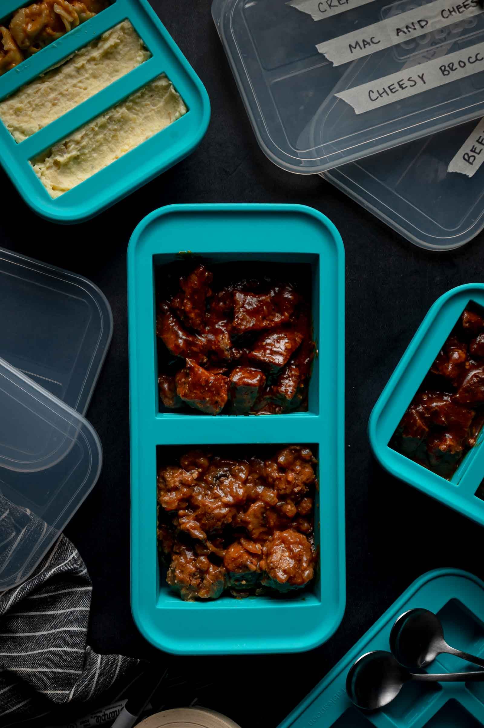 Blue meal prep containers filled with cooked food are arranged on a dark surface, with clear plastic lids nearby.