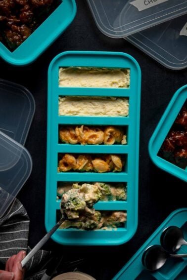 A teal tray with five sections holds various side dishes, including macaroni and cheese and cheesy broccoli, with a spoon serving food into one compartment. Other covered containers are nearby.