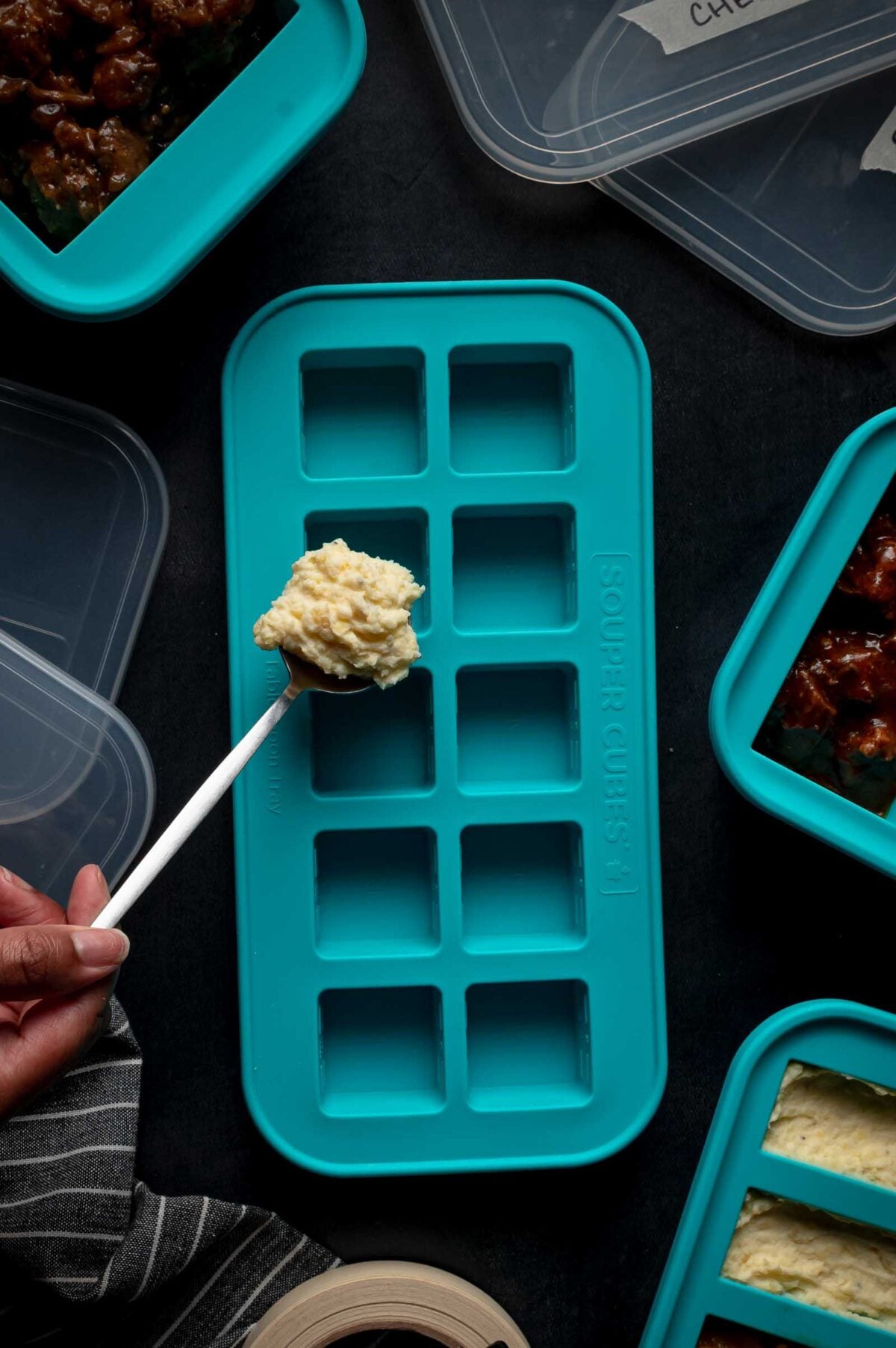 A hand holds a spoon of mashed potatoes over a blue silicone freezer tray surrounded by other filled trays on a dark surface.