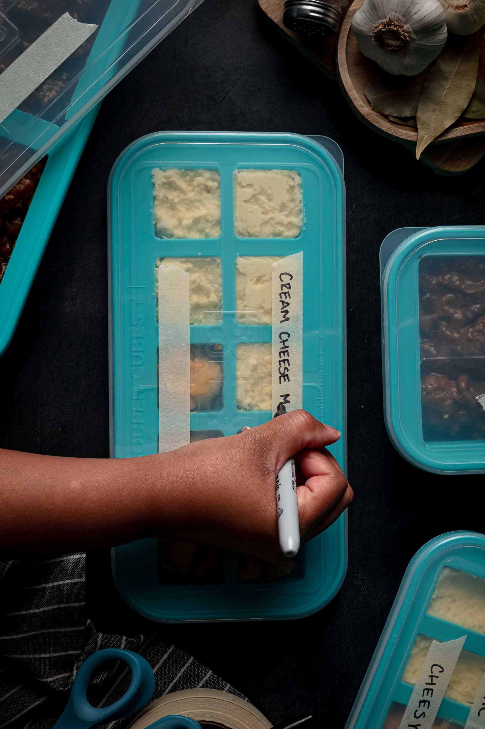 A hand labels a blue silicone freezer tray filled with cream cheese mashed potatoes using a black marker. Other labeled containers and kitchen items are nearby on a dark surface.