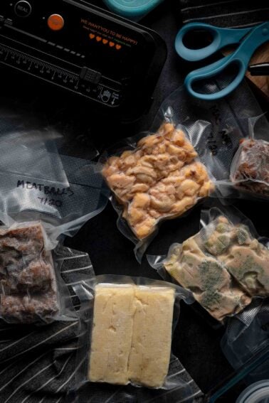 Vacuum-sealed bags filled with different foods are arranged on a dark surface near a vacuum sealer, scissors, and striped cloth.