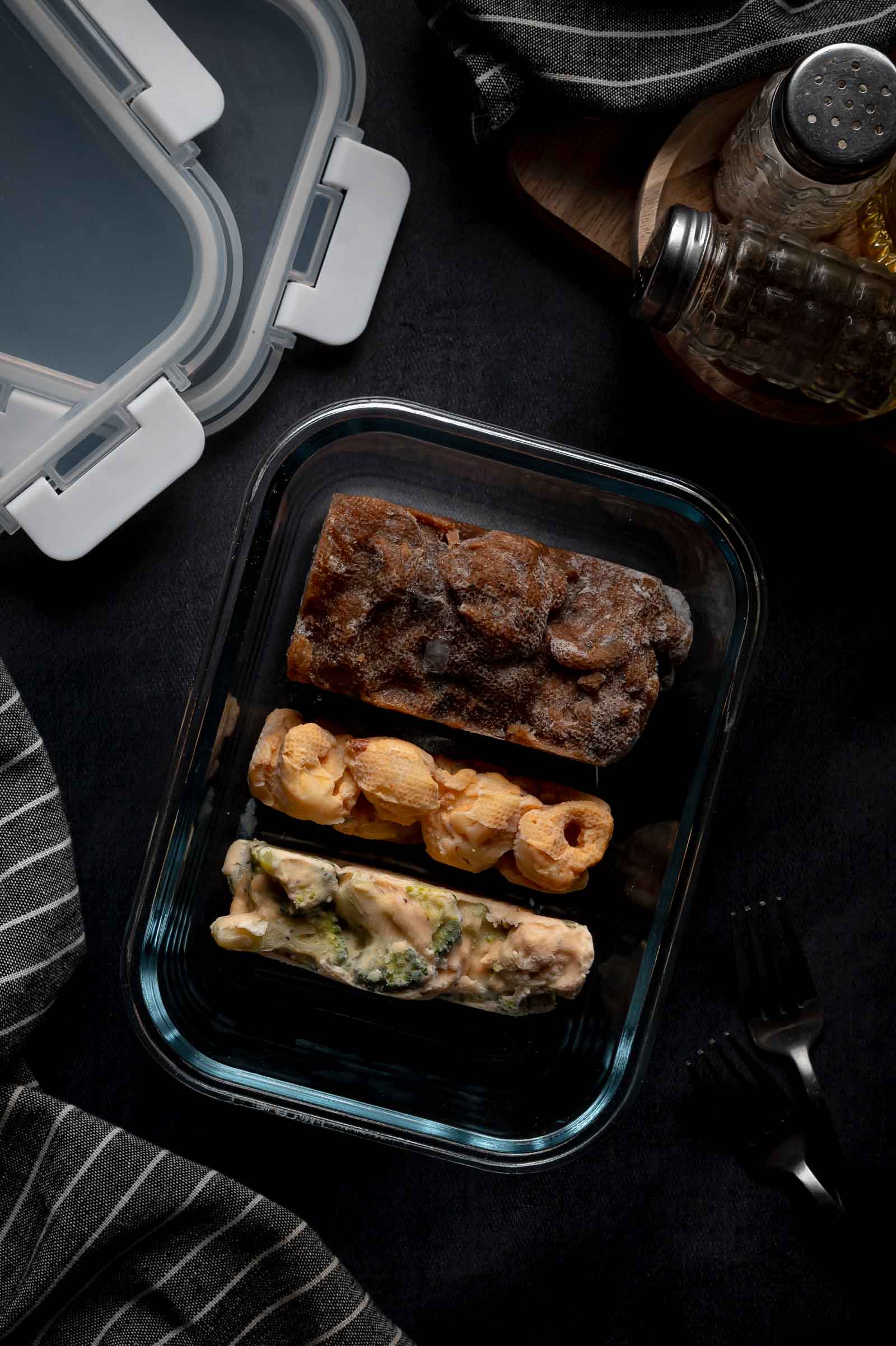 Three different frozen meal prep cubes are placed in a rectangular glass container on a dark table with a striped towel, black fork, and seasoning shaker nearby.