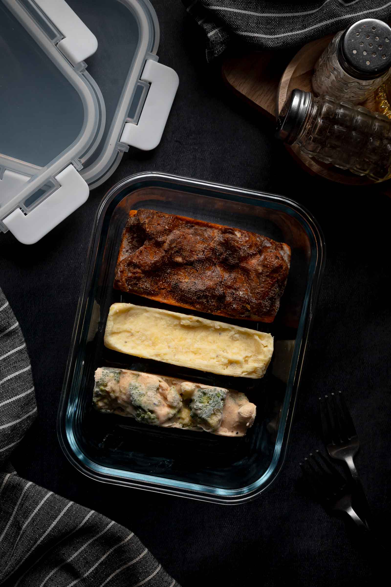 Glass container with three frozen food portions: one beef tips, one mashed potatoes, and one cheesy broccoli cube, on a dark surface with utensils and seasonings nearby.