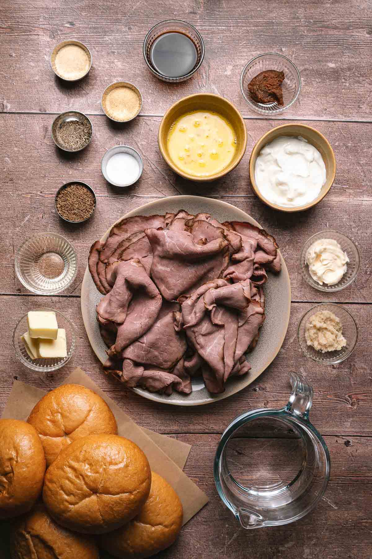 A plate of sliced roast beef is surrounded by sandwich buns, seasonings, sauces, butter, and a measuring cup on a wooden surface.