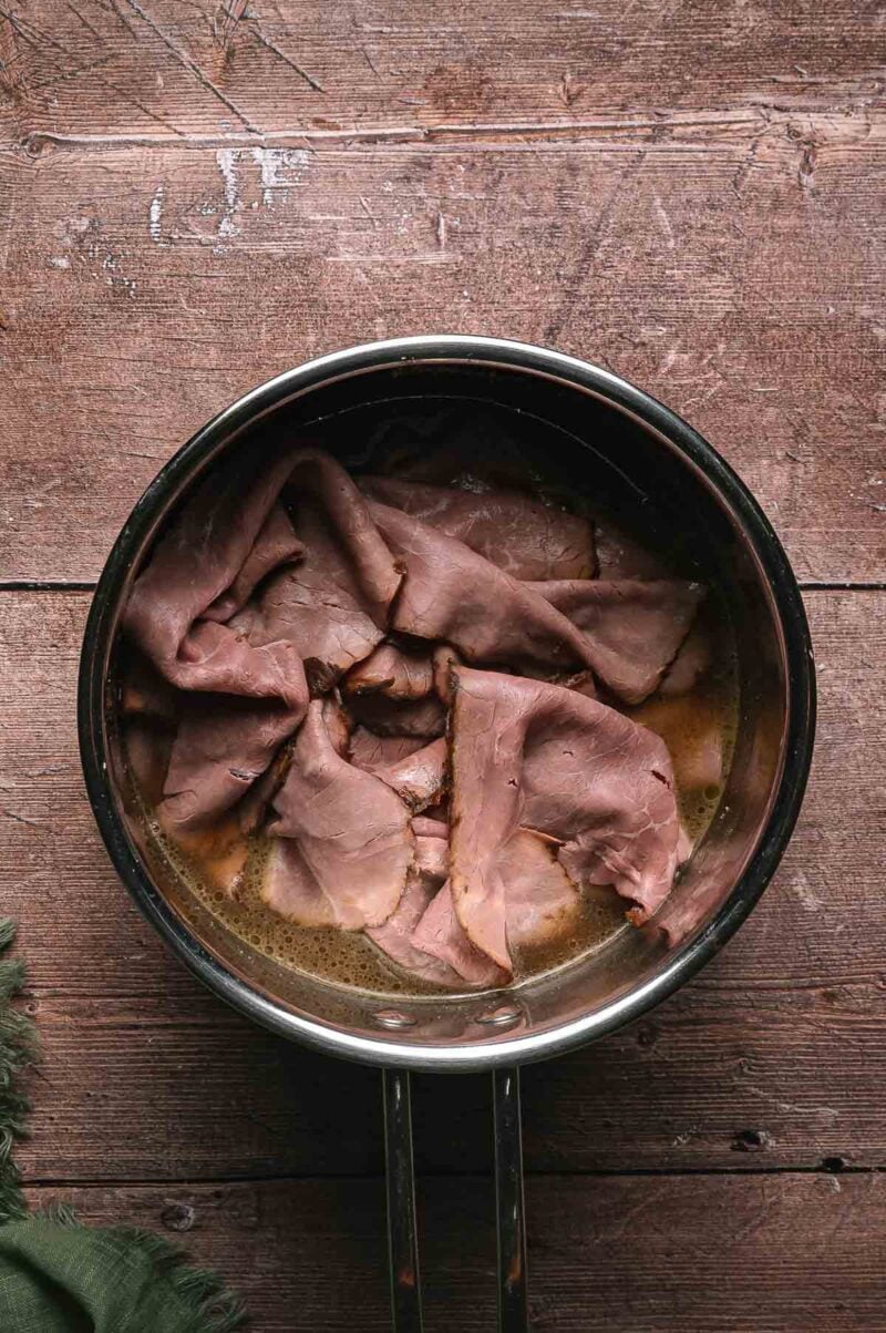 Sliced roast beef sits in au jus inside a metal saucepan placed on a rustic wooden surface.