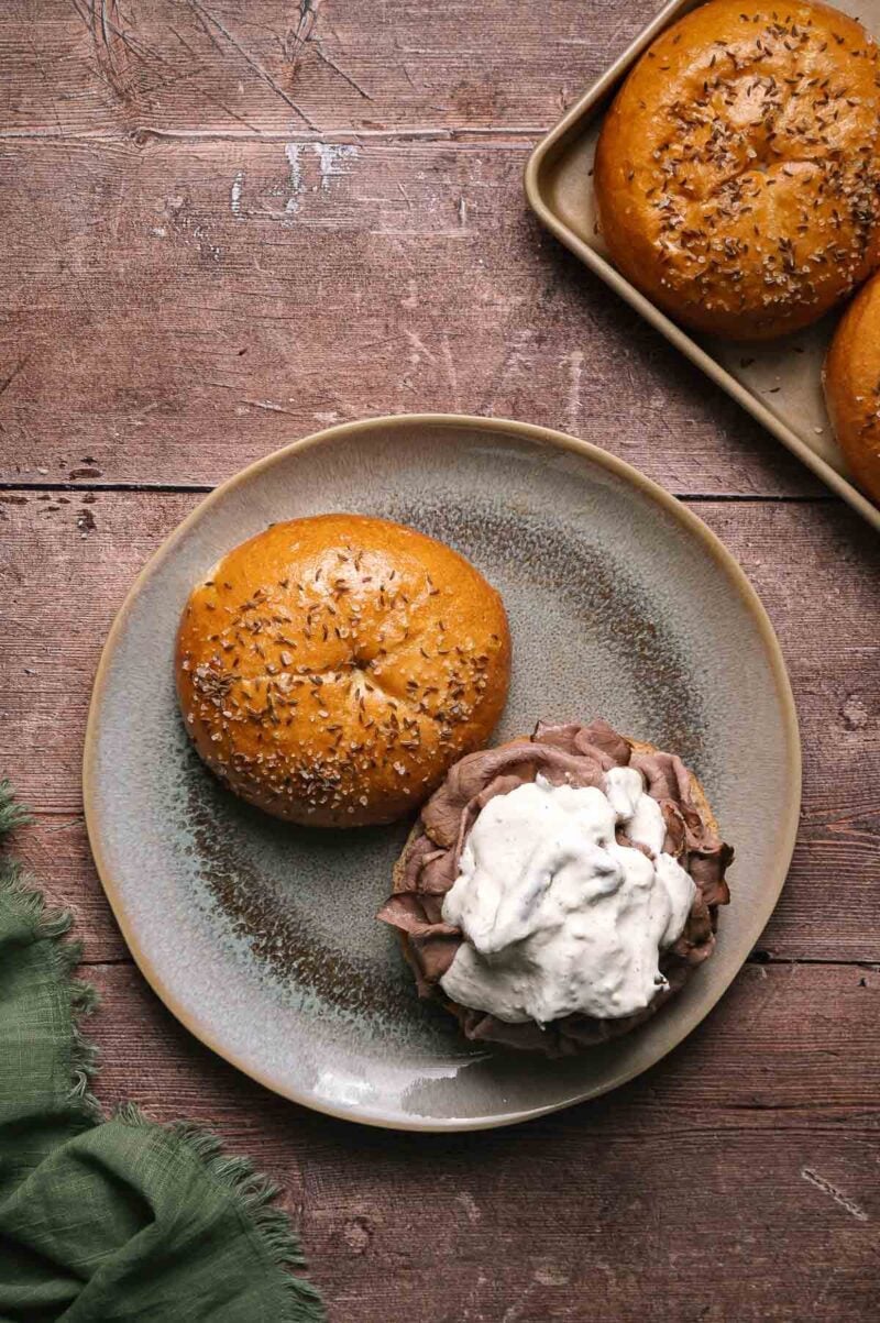 A sandwich with sliced roast beef and a dollop of horseradish sauce on a plate beside a salted bun, with additional buns on a tray in the background.