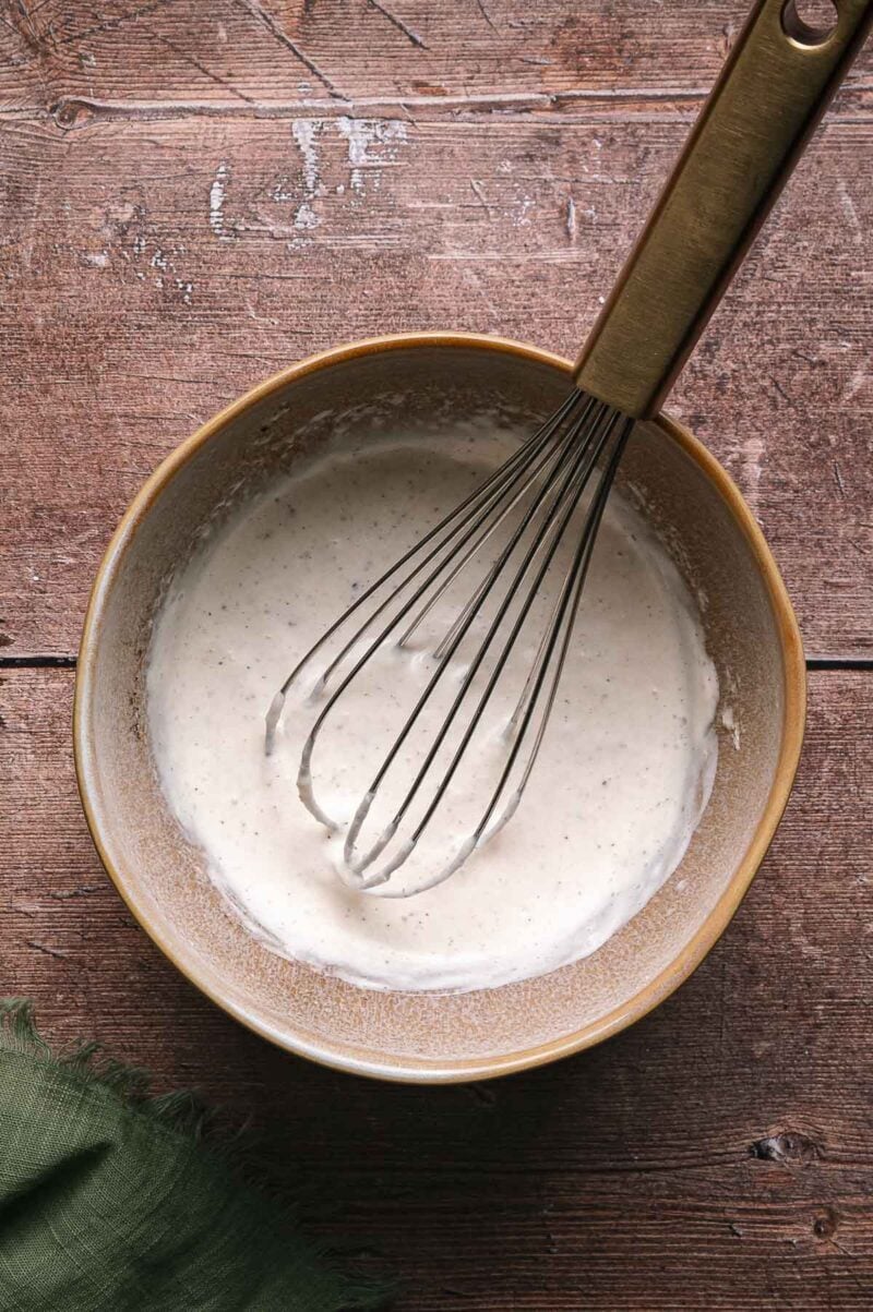 A whisk rests in a bowl of creamy, light-colored horseradish sauce or batter on a wooden surface with a green cloth nearby.