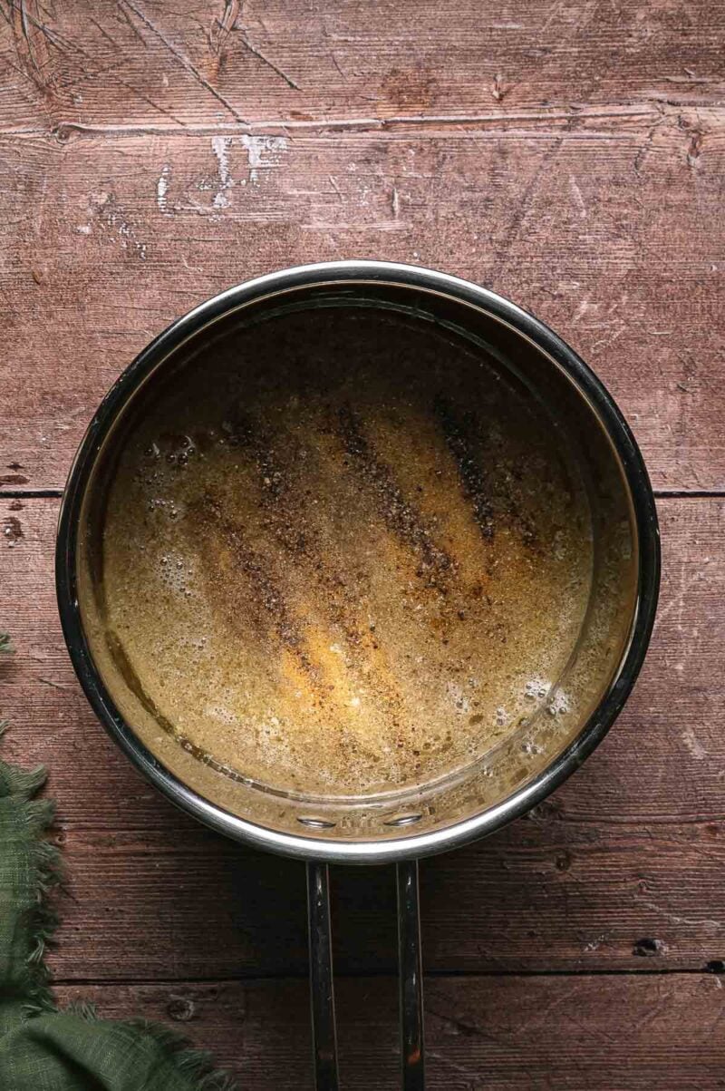 A saucepan containing au jus with spices sits on a wooden surface, with visible bubbles and brown specks in the liquid.