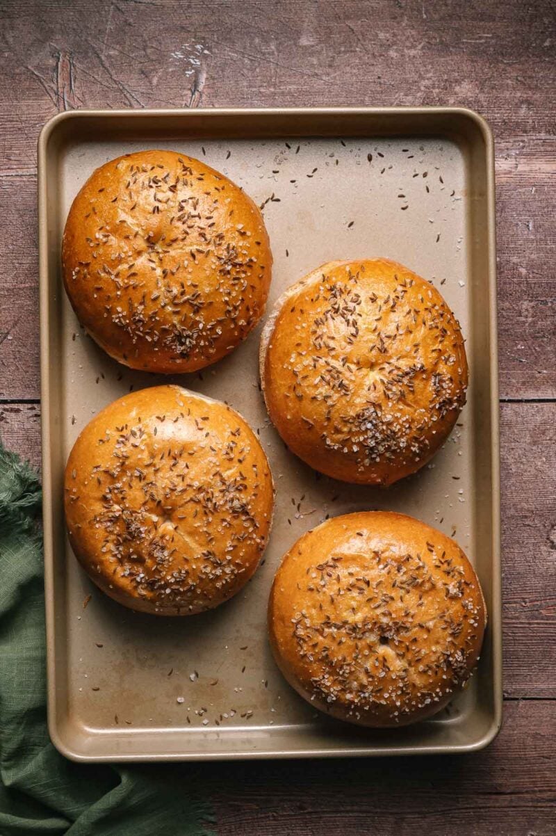 Four round sandwich buns topped with caraway seeds and coarse salt are arranged on a baking sheet on a wooden surface.