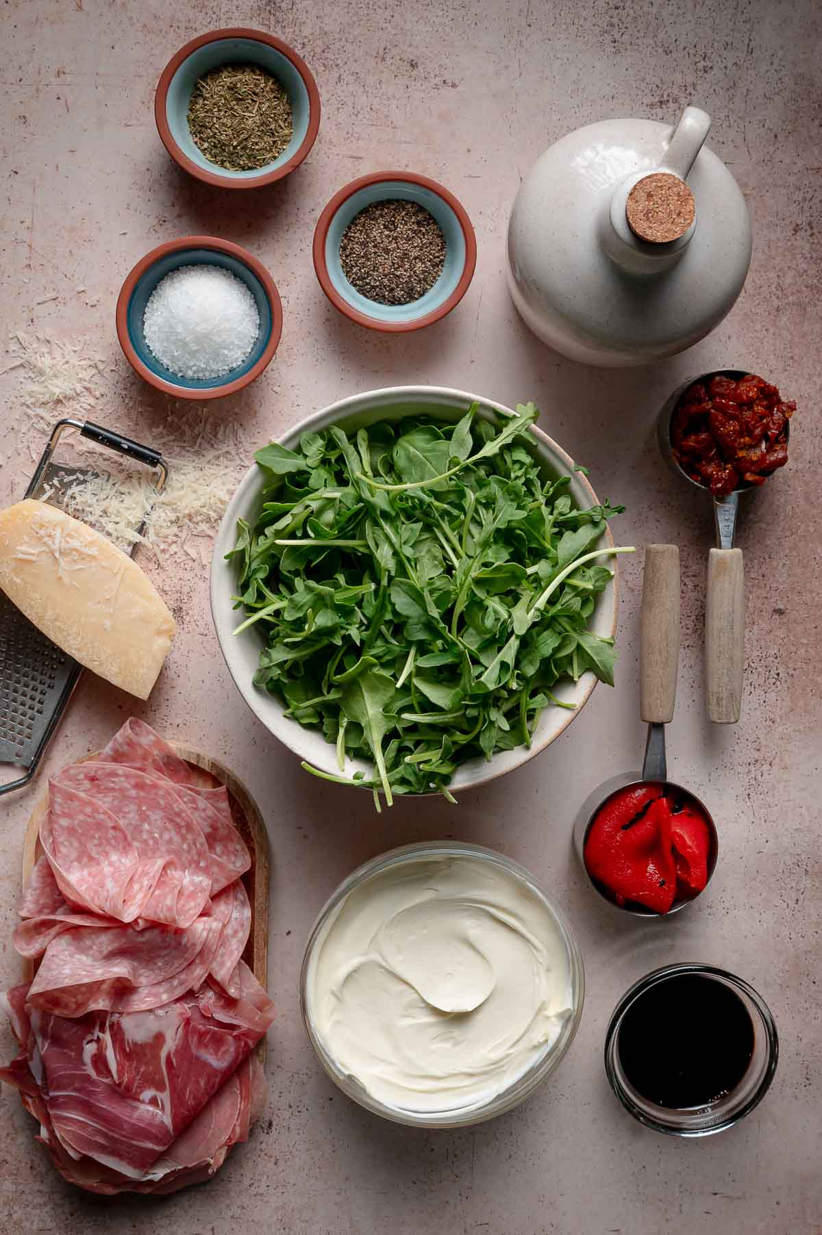 A variety of ingredients including arugula, cured meats, cheese, cream, spices, roasted peppers, sun-dried tomatoes, vinegar, and oil are arranged on a countertop.