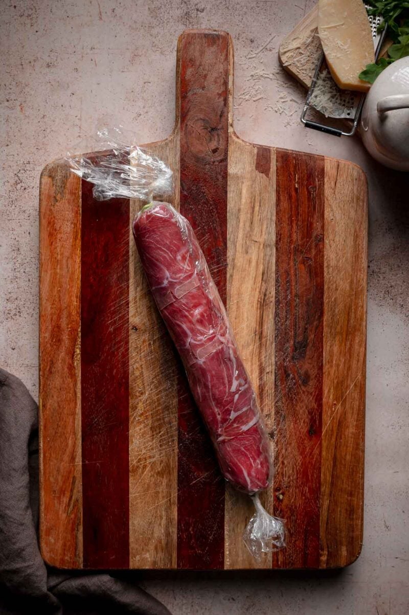 Italian sushi wrapped in a tight log in cling wrap is placed on a wooden cutting board.