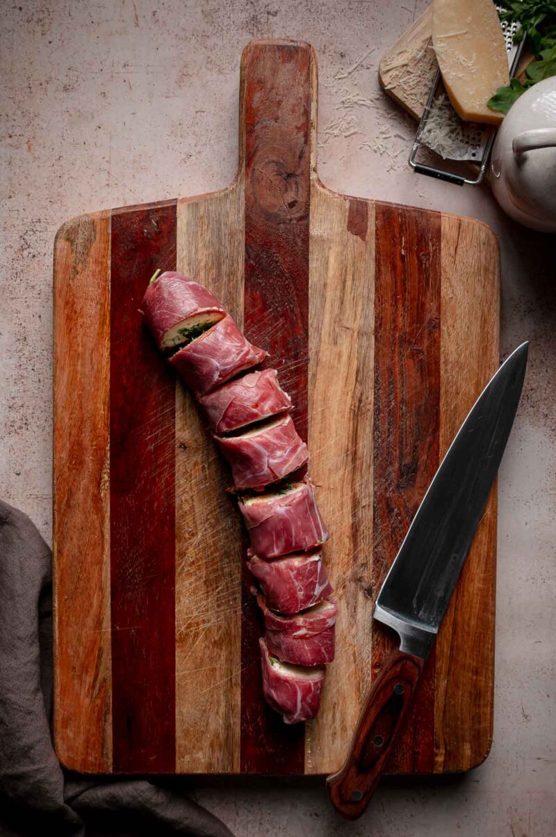 Sliced prosciutto-wrapped Italian sushi roll on a wooden cutting board next to a large knife.