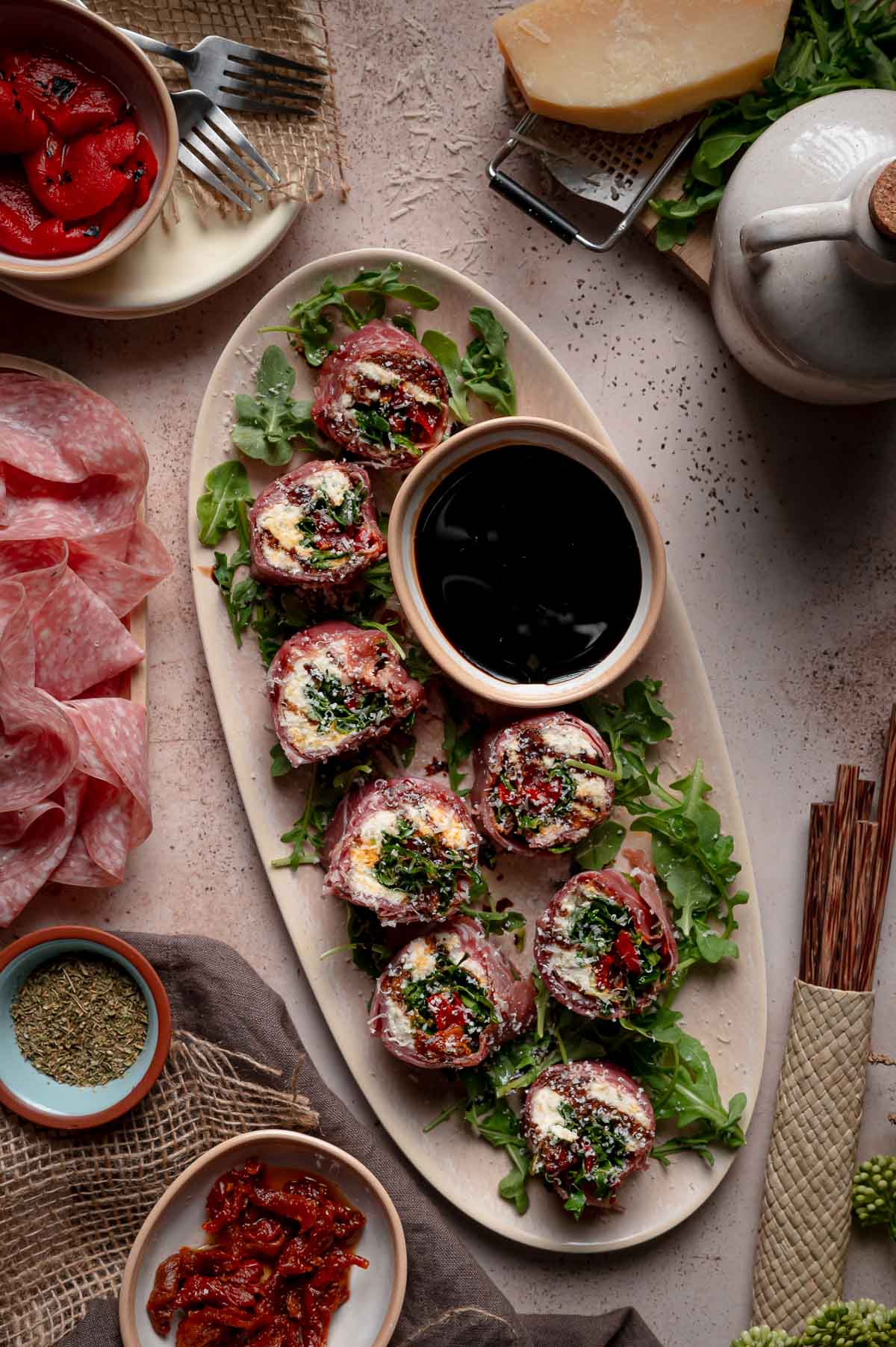 An oval platter with prosciutto-wrapped italian sushi rolls on greens, served with a bowl of balsamic oil for dipping; surrounding dishes include meats, sun-dried tomatoes, and seasonings.