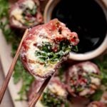Chopsticks hold a piece of prosciutto-wrapped italian sushi filled with greens and cheese over a plate with more appetizers and a small bowl of dark dipping sauce.