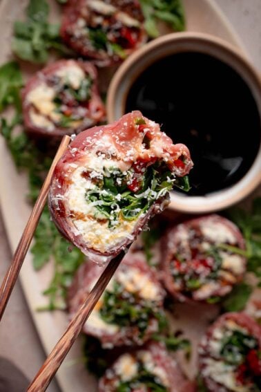 Chopsticks hold a piece of prosciutto-wrapped italian sushi filled with greens and cheese over a plate with more appetizers and a small bowl of dark dipping sauce.